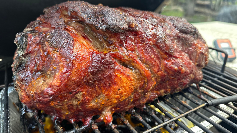 A pork shoulder, with a nice charred crust, on a grill with a drip pan underneath and a thermometer in the background