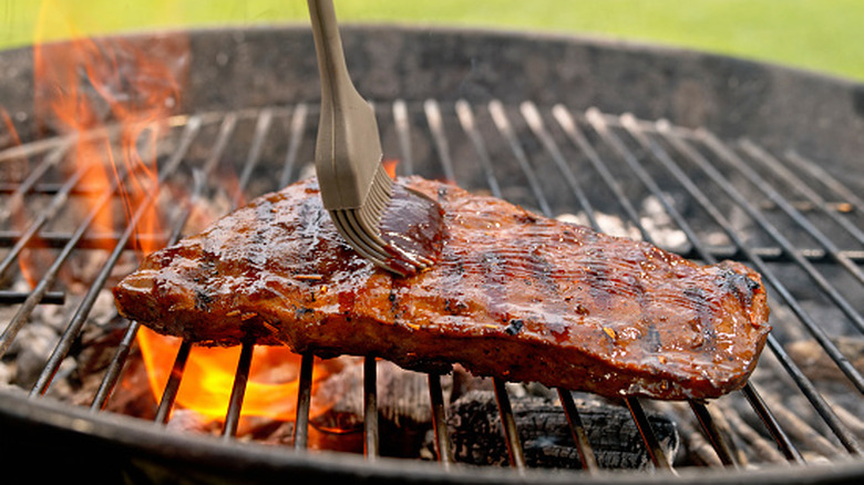 A rack of ribs being sauced on a kettle grill with flames coming up on one side