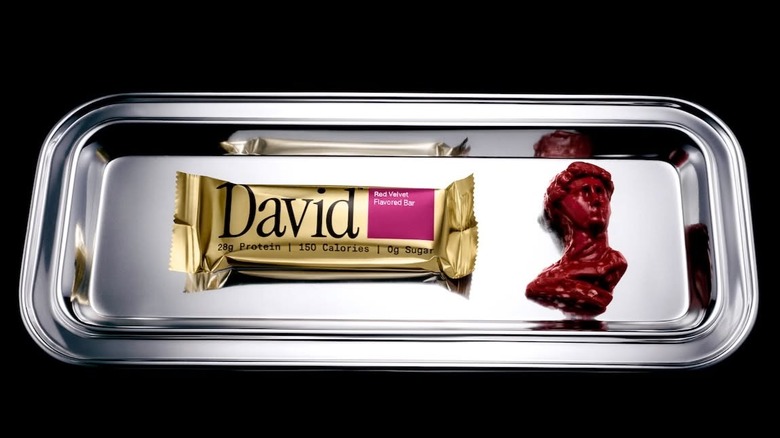 A David protein bar placed on a silver platter with a small greek statue replica against a black background.
