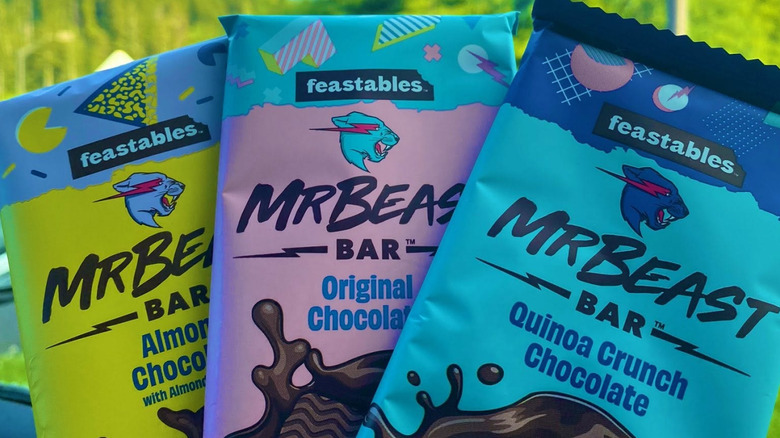 A hand holding a selection of three Mr Beast Feastable bars in a car against a green background.