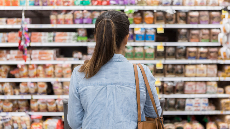 A woman standing with a shopping cart in front of a shelf full of food in the bread aisle of a grocery store.