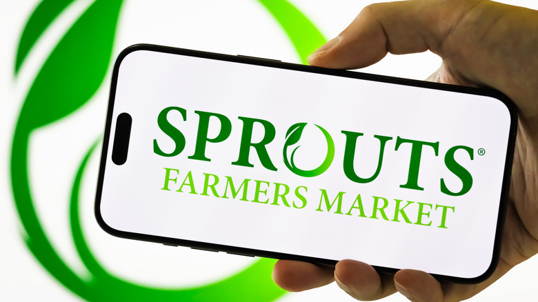 Hand holding phone with Sprouts logo in front of logo on white background