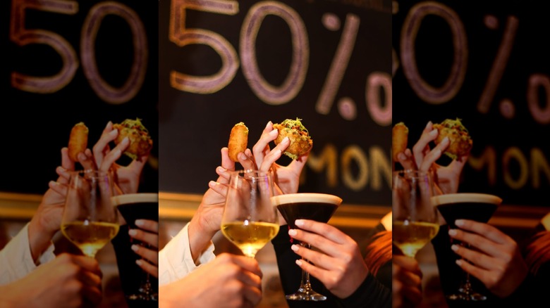 Hands toasting drinks while eating appetizers in front of 50% off Monday sign at Neme Gastro Bar