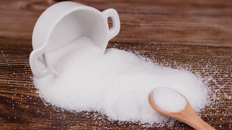 A ceramic sugar container tipped over, spilling sugar on a wooden table. A wooden spoon filled with sugar rests nearby, creating a rustic, natural scene.