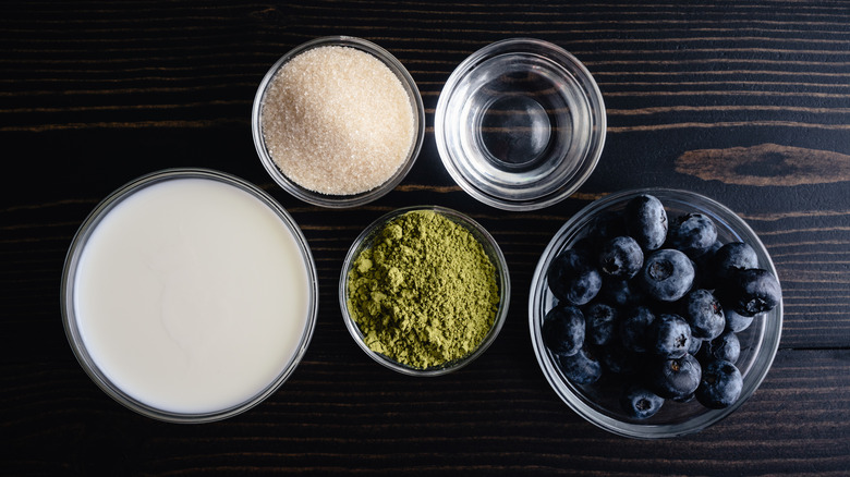 Blueberry Matcha Latte Ingredients on a Dark Wooden Table: Green tea powder, fresh blueberries, organic sugar, milk, and water in glass prep bowls.