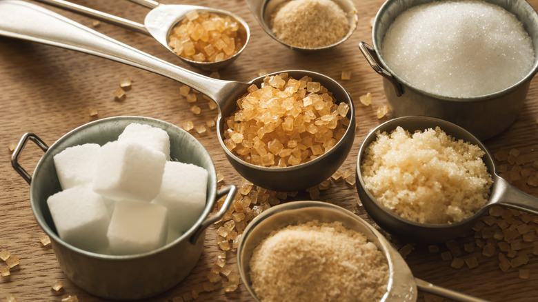 Assortment of Sugar (From Left to Right: White Crystal Sugar, Brown Sugar Cubes, White Sugar Cubes, Brown Sugar Syrup, Brown Rock Sugar, Brown Granulated Sugar, White Granulated Sugar) in Bowls on Brown Background Directly Above View.