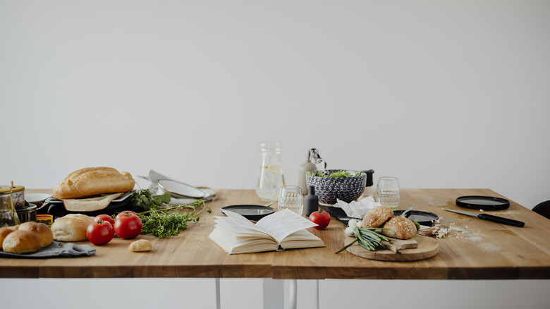 Messy dining table with scattered ingredients, crumbs and an open book against a white wall