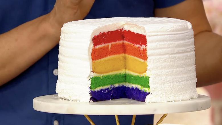 A rainbow cake with various colorful layers coated in whipped cream
