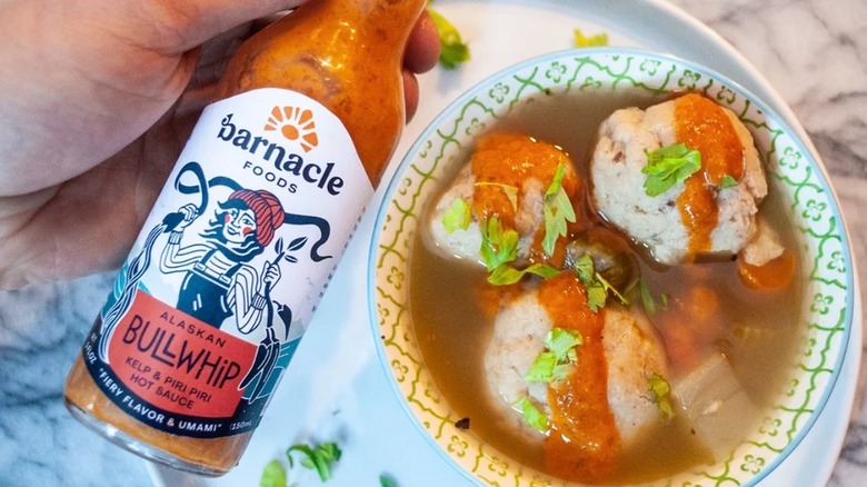 Hand holding bottle of Barnacle Bullwhip Hot Sauce over bowl of meatball soup