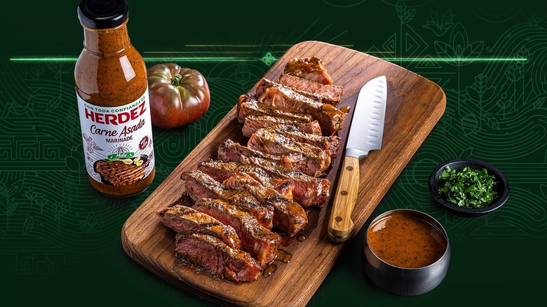 Bottle of Herdez Carne Asada marinade next to sliced steak on cutting board, all on green background