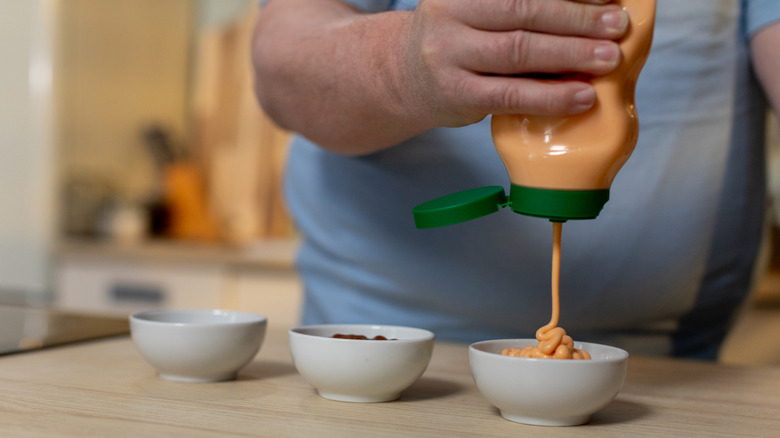 Hand squeezing bottle of sauce into white cup on counter