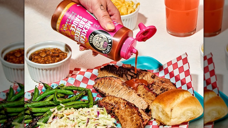 Hand pouring Tia Lupita Mexican Barbecue Sauce onto grilled meat with coleslaw, roll, green beans, baked beans beside