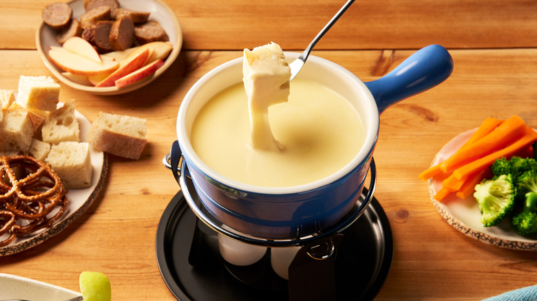 A person dipping bread in a cheese fondue in the center with various ingredients around it