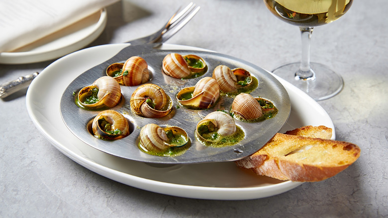 A plate of escargot served in the shell on escargot baking plate with crusty bread and a glass of wine in the back