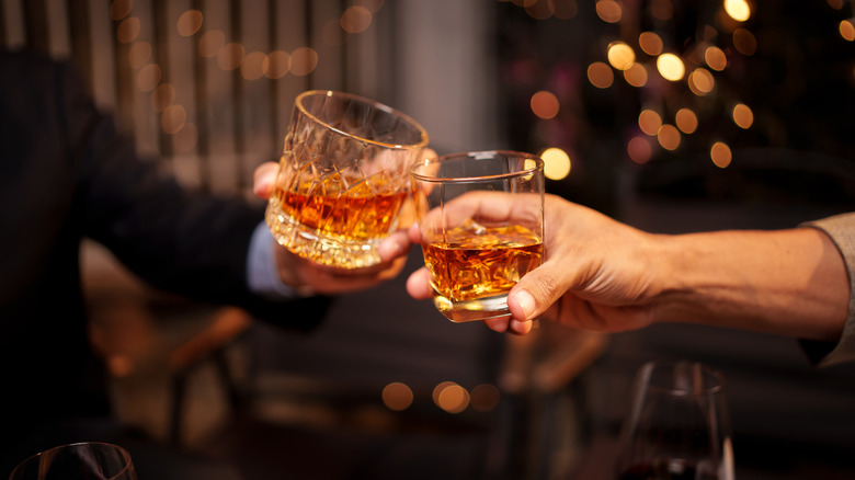 Two people clinking two glasses of whiskey together with twinkling lights in the background