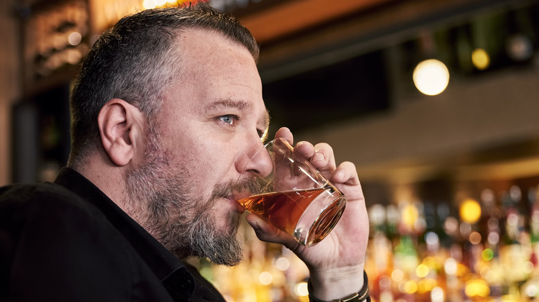 A close up of a person drinking whiskey from a tumbler with a blurred bar in the background