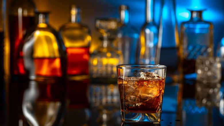 A whiskey glass in the foreground with a wide selection of unbranded bottles in the background
