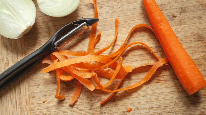 Carrot and peels next to vegetable peeler on wooden cutting board