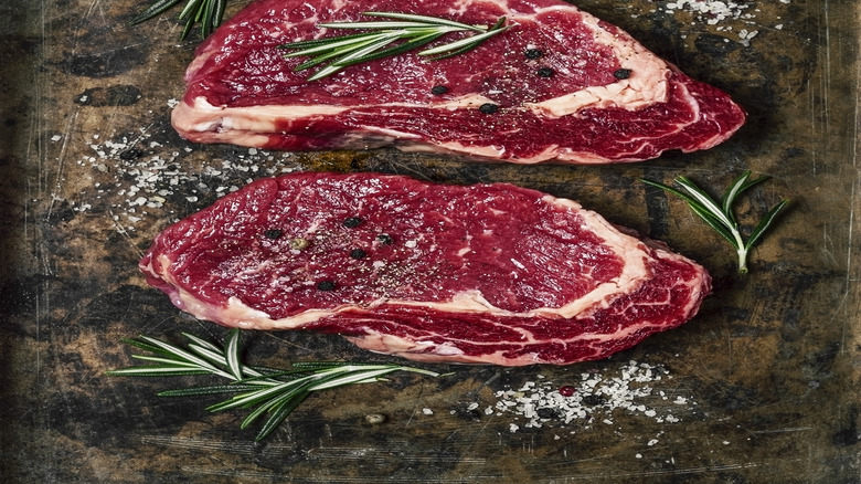 Raw ribeye steaks on a tray with coarse salt, peppercorns, and rosemary