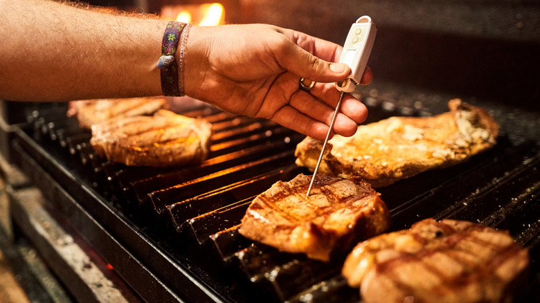 A hand holding a meat thermometer checking the temperature of steaks on a grill