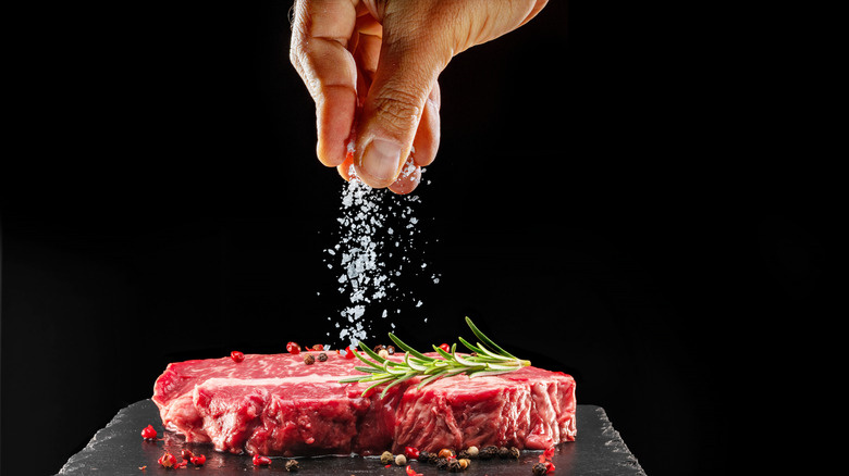 A hand sprinkling salt onto a raw ribeye steak