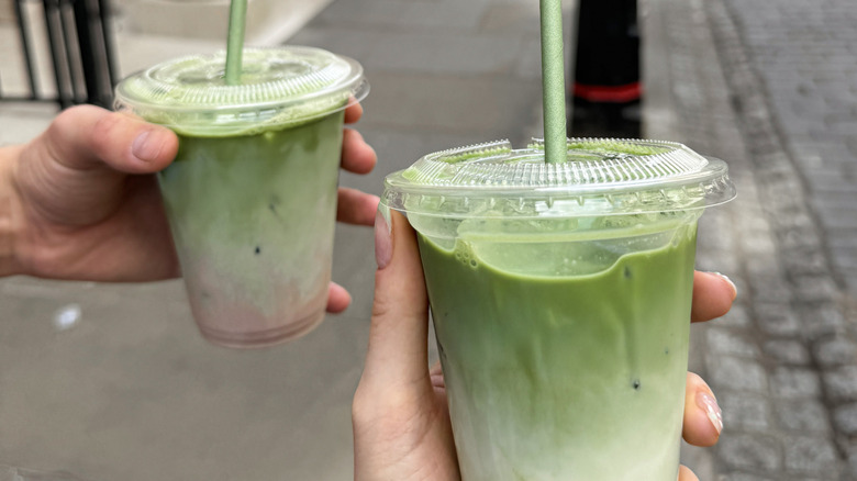Two hands holding matcha lattes in takeaway cups