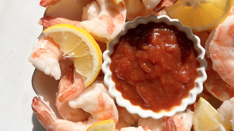 Cocktail sauce in white bowl surrounded by shrimp and cut lemons