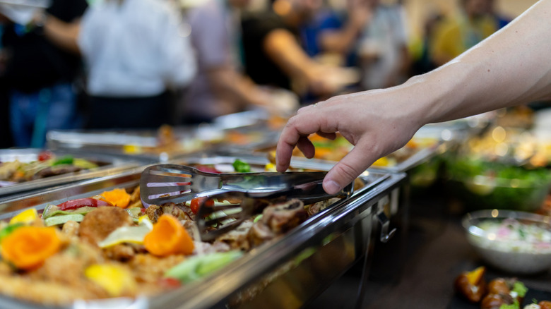 A hand reaches out to grab a serving of assorted dishes from a buffet, surrounded by many people at a busy gathering.