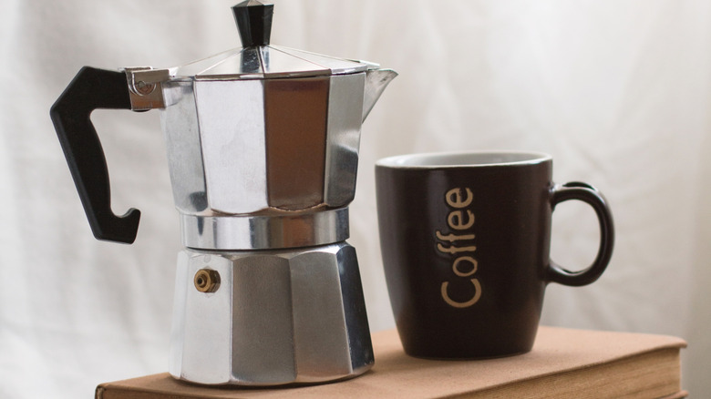A moka pot and a mug that reads, "coffee" sitting on a book.