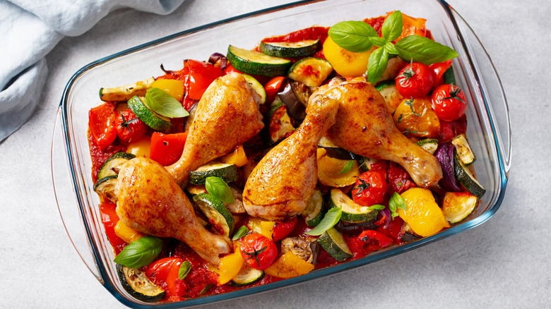 Chicken drumsticks and roasted vegetables in glass baking dish with fresh basil used as garnish