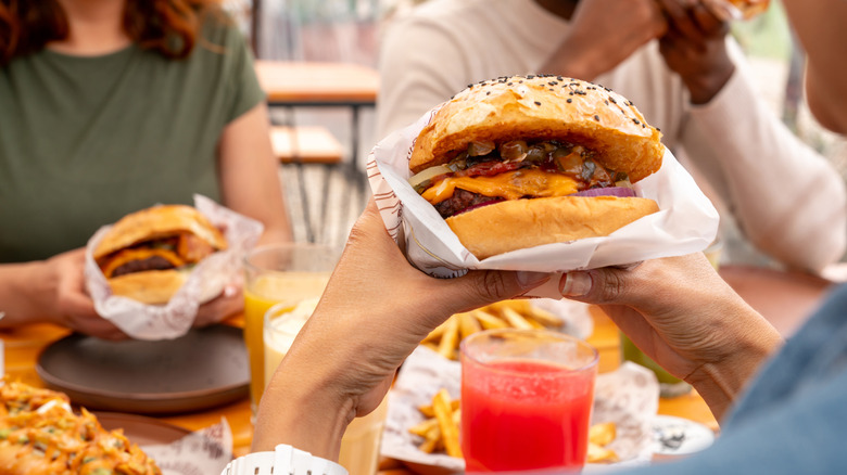 Three people sitting down and holding fast food burgers with other items like fries and hot dogs on table