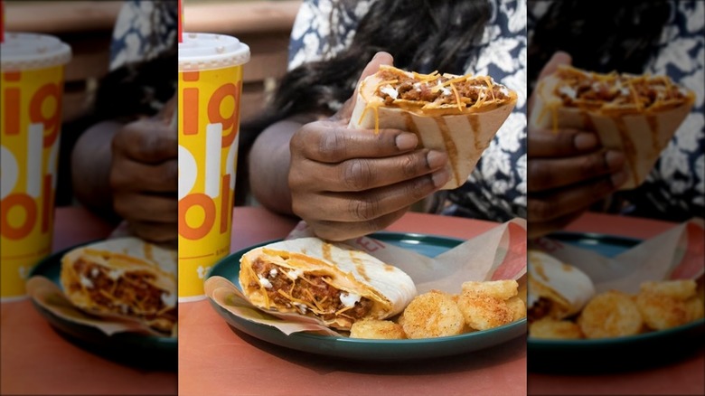 Customer holding Taco John's Stuffed Grilled Taco half above plate holding other taco half and fried potatoes