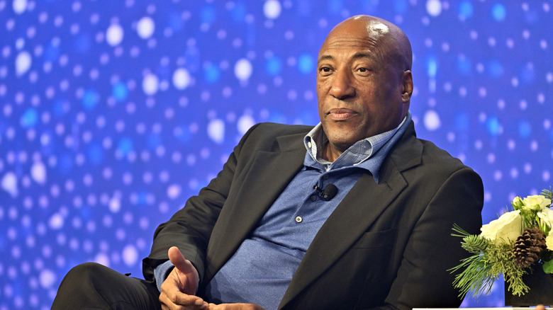 Byron Allen wearing a suit and sitting onstage in front of blue background