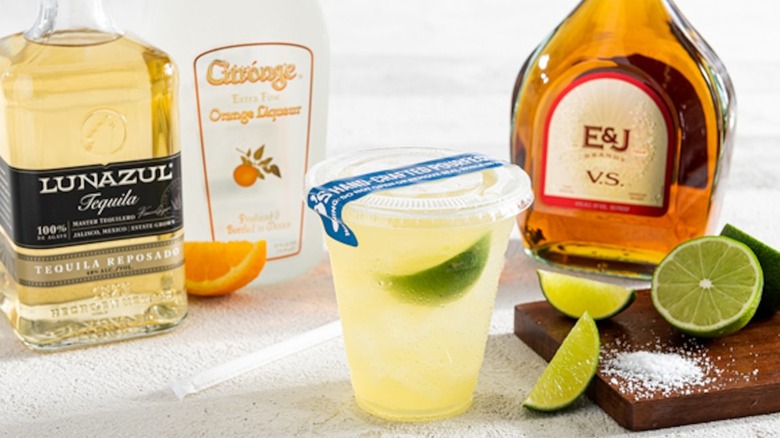 Chili's to-go Presidente Margarita in a plastic cup with tequila, orange liqueur, and E&J bottles in the background, limes with salt on a cutting board, and an orange slice against a white table background