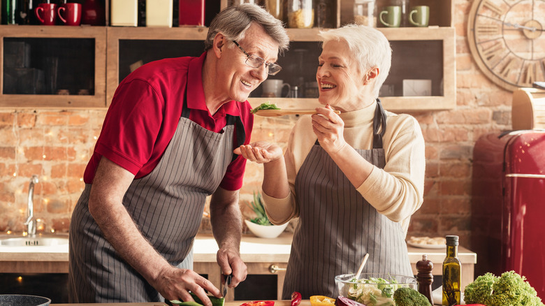 Grey-haired woman getting grey-haired man to taste food on a spoon in kitchen