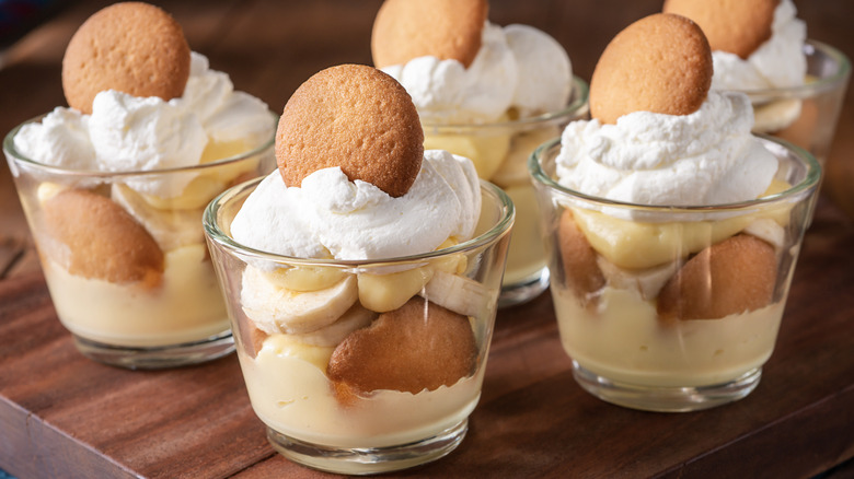 Glasses of banana pudding on wooden cutting board, with vanilla wafers and whipped cream