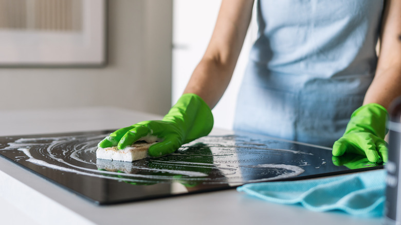 Person wearing green gloves, cleaning stovetop with a sponge