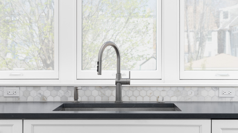 Kitchen sink detail with white cabinets, grey countertop, a marble hexagon backsplash and large windows.