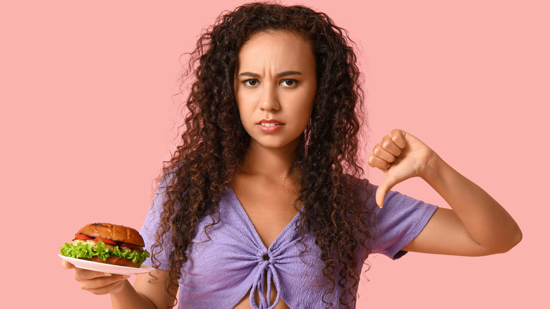 Woman in a purple shirt holding a burger, looking annoyed, and giving a thumb's down