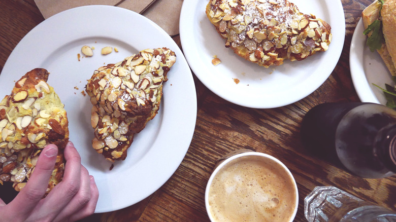 Hand picking up an almond croissant from a plate on a table laden with coffee, water, and a sandwich