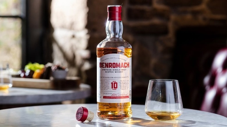An opened bottle of Benromach 10-year shown on a table next to a filled glass of whisky