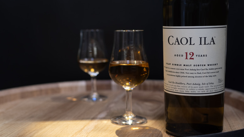 A bottle of Caol Ila 12-year perched on top of a barrel with two full glasses shown in the background
