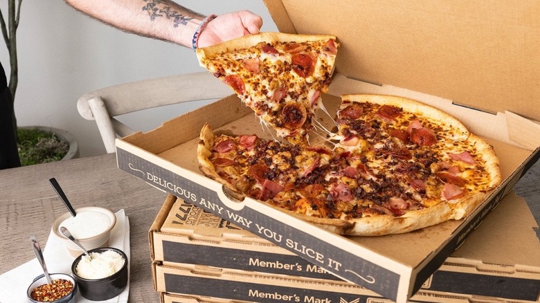A hand reaching for a slice of Sam's Club Four-Meat pizza from an open box on a stack of pizza boxes