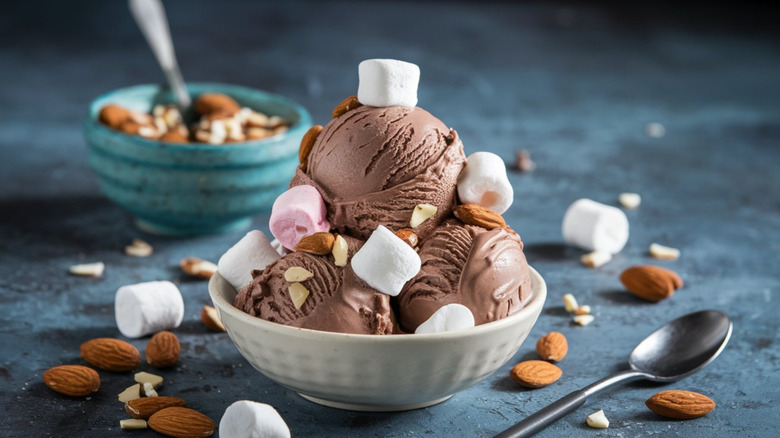Scoops of rocky road ice cream in white bowl with almonds and marshmallows scattered around it
