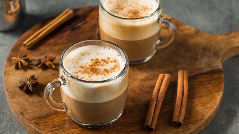 Two glass mugs filled with dirty chai latte on a wooden board surrounded by anise and cinnamon sticks