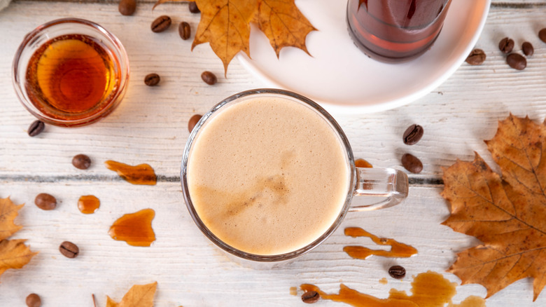 Top view of a maple cappuccino with pumpkin spice surrounded by maple syrup, coffee beans, and maple leaves