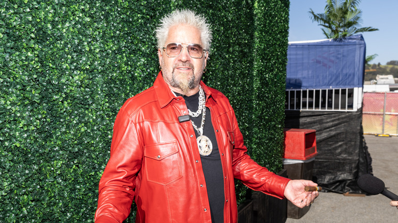Guy Fieri wearing red leather jacket posing against grass wall