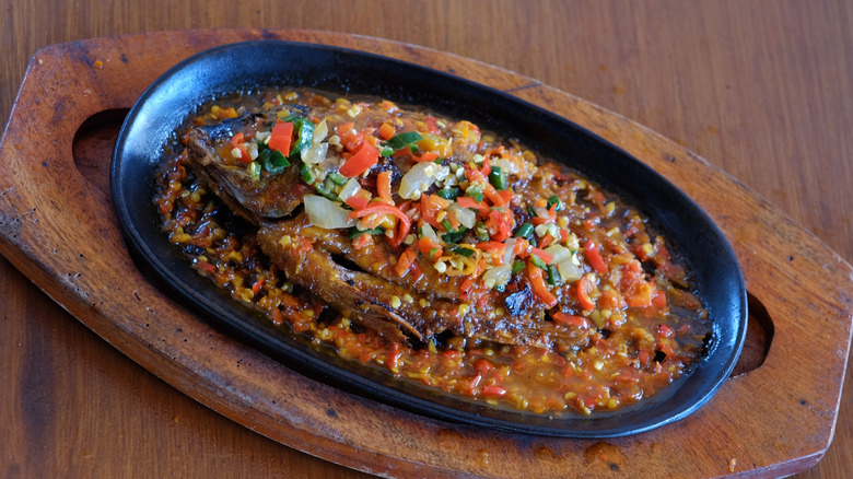 Fish drowning in sauce and chopped veggies in black oblong dish.