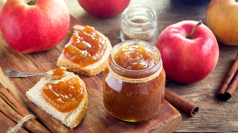 A jar of homemade apple butter surrounded by toast, apples, and cinnamon sticks
