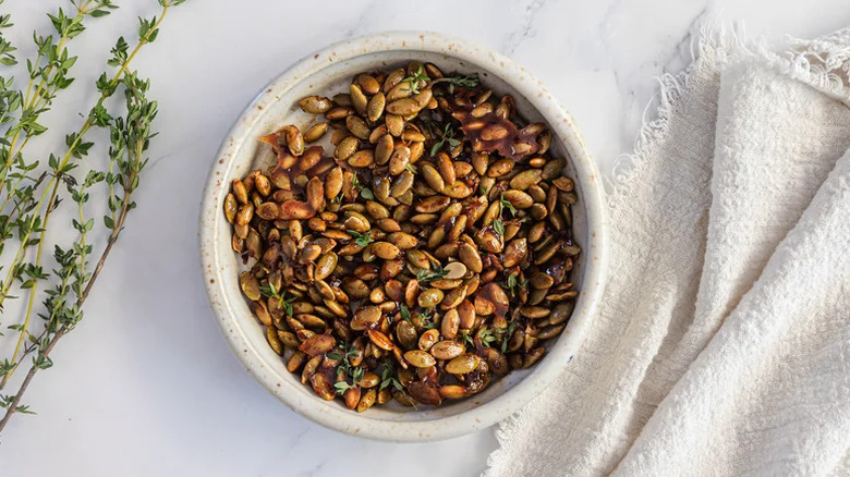 A bowl full of maple roasted pumpkin seeds next to thyme sprig and linen cloth